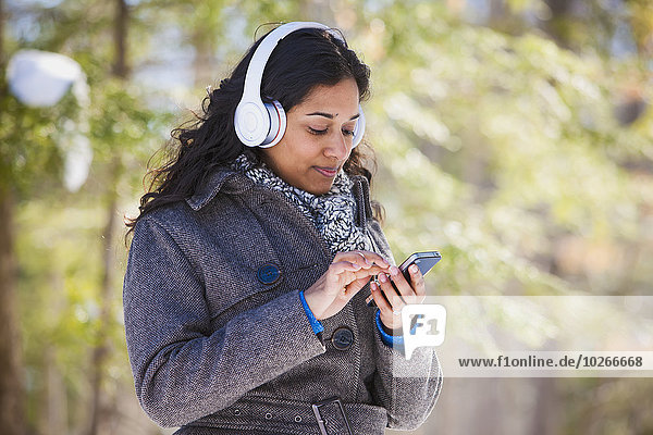 Young woman of South Asian ethnicity using cell phone and Bluetooth headphones in Scanlon Creek Conservation Area; Ontario  Canada