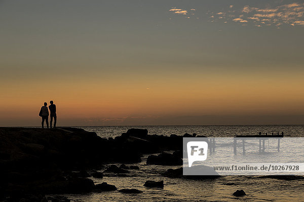 Silhouette of two people standing at the water's edge watching the sunset over the water; Paphos  Cyprus