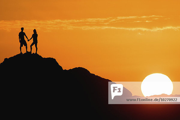 Silhouetted couple holding hands on a big rock at sunset; Oahu  Hawaii  United States of America