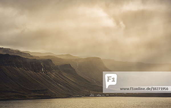 Lichtstrahlen scheinen auf die Berge und das verlassene Dorf Djupavik in Island.