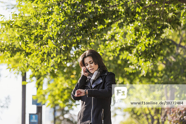Ältere Geschäftsfrau  die mit ihrem Handy telefoniert und auf die Uhrzeit achtet  während sie in einem städtischen Park im Herbst spazieren geht; Edmonton  Alberta  Kanada