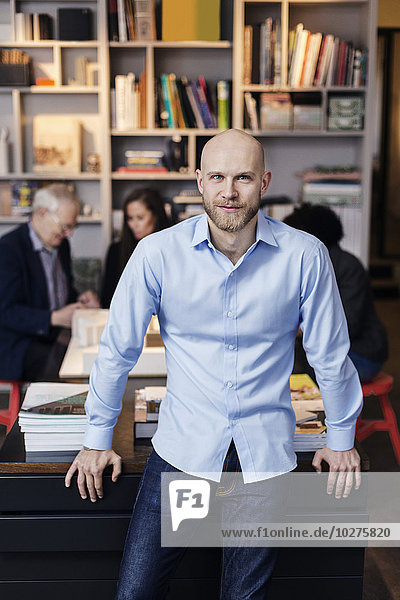 Porträt eines selbstbewussten Geschäftsmannes  der sich auf den Schreibtisch lehnt  während seine Kollegen im Hintergrund arbeiten.