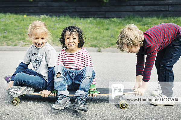 Porträt von glücklichen Kindern  die auf dem Skateboard sitzen  während ein Freund es auf dem Hof zieht.