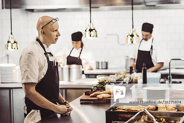 Side view of male chef standing at kitchen counter with colleagues working in background