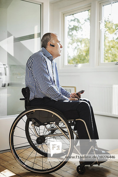 Mature man holding mobile phone while using Bluetooth in wheelchair at home