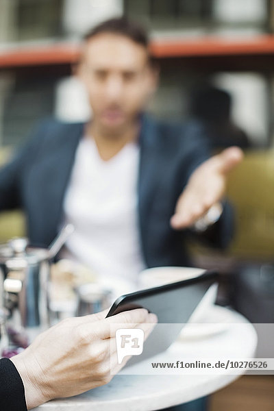 Geschäftsmann gestikuliert gegenüber einer Frau  die ein digitales Tablett im Straßencafé hält.