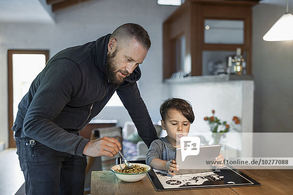 Vater füttert Sohn mit digitalem Tablett am Tisch im Haus