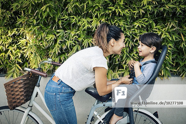 Glückliche Frau schaut auf den Sohn  der auf dem Rücksitz des Fahrrads gegen die Pflanzen sitzt.