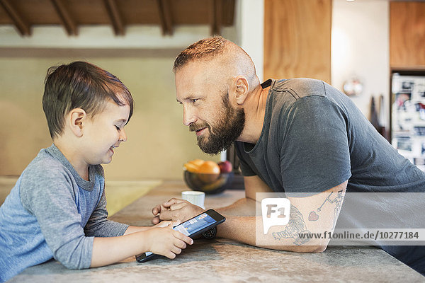 Vater sieht lächelnden Sohn mit digitalem Tablett am Tresen in der Küche.