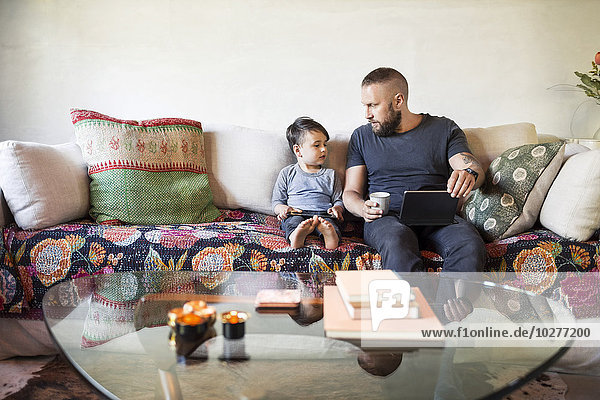 Vater und Sohn mit digitalen Tabletts sitzen zu Hause auf dem Sofa