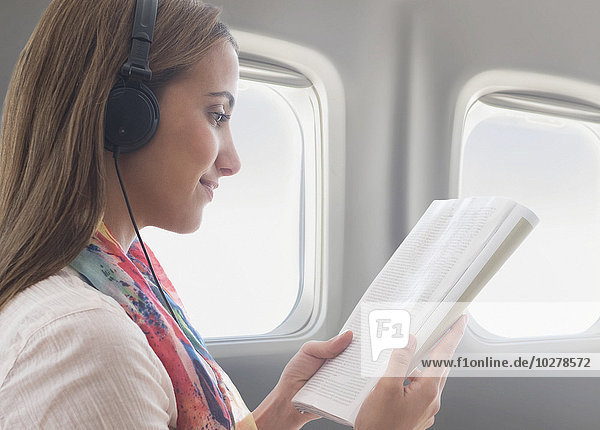 Junge Frau trägt Kopfhörer beim Lesen eines Buches im Flugzeug
