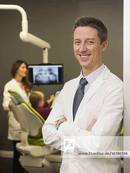 Porträt eines Zahnarztes mit einem Kollegen und einem Patienten (8-9) im Hintergrund