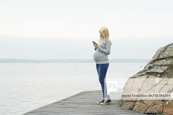 Schwangere Frau auf dem Bootssteg  die ein Mobiltelefon benutzt