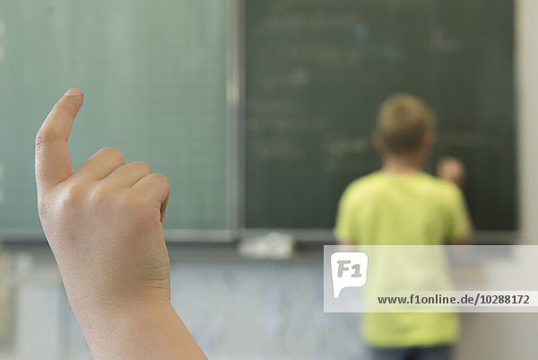 Ein Schüler schreibt an der Tafel und eine Schülerin hebt die Hand in einem Klassenzimmer  München  Bayern  Deutschland