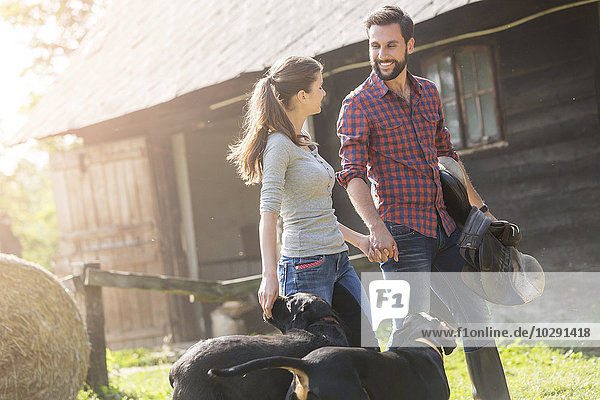 Couple with saddle and dogs holding hands outside rural barn