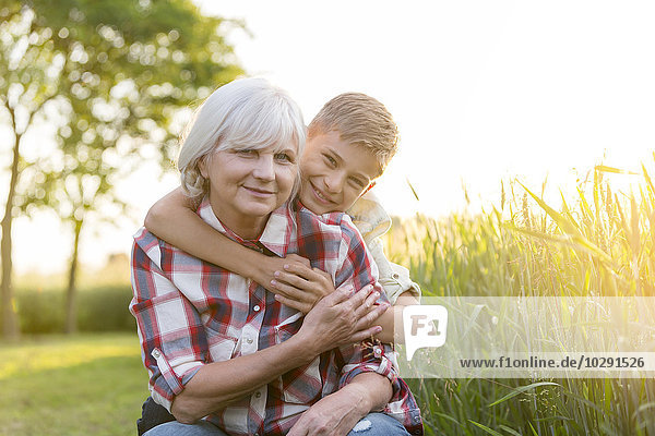 Portrait lächelnde Großmutter und Enkel  die sich in der Nähe des sonnigen ländlichen Weizenfeldes umarmen.