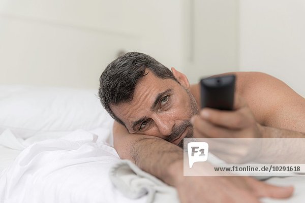 Nackt geschulterterter reifer Mann auf dem Bett liegend mit Fernbedienung
