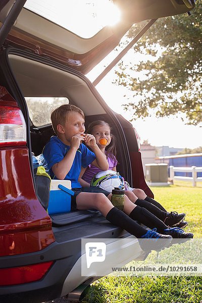 Junge und jüngere Schwester sitzen im Kofferraum und essen Orangen in der Fußballtrainingspause.
