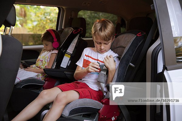 Girl using digital tablet whilst brother fastens seat belt in car back seat