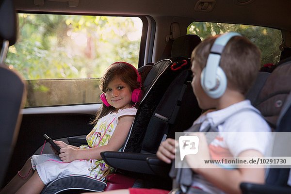 Junge und jüngere Schwester tragen Kopfhörer und benutzen ein digitales Tablett auf dem Rücksitz des Autos.