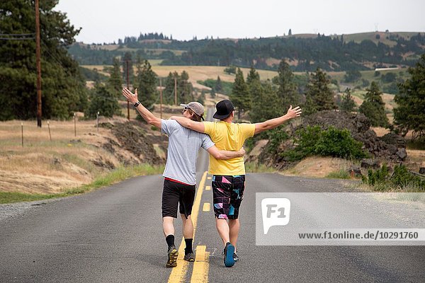 Two men walking along road together  arms raised  appreciating view  rear view
