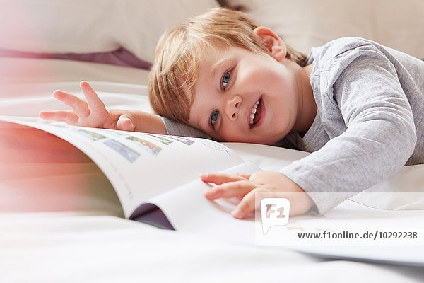 Head and shoulders of boy on bed lying on front holding storybook  looking away smiling