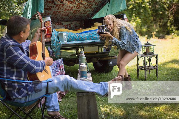 Young woman photographing boyfriend playing acoustic guitar whilst camping