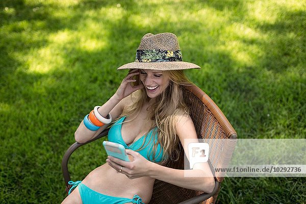 Junge Frau im Bikini und Sonnenhut beim Lesen von Smartphone-Texten im Garten