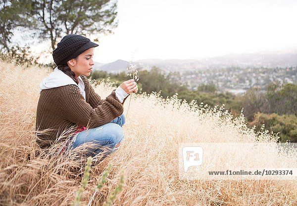 Young woman crouching on grassy hill holding dry blade of grass