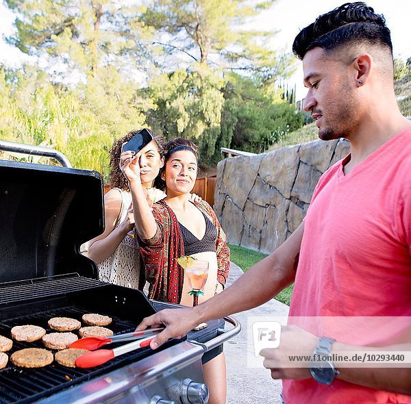 Young man cooking on barbecue in front of sisters taking smartphone selfie