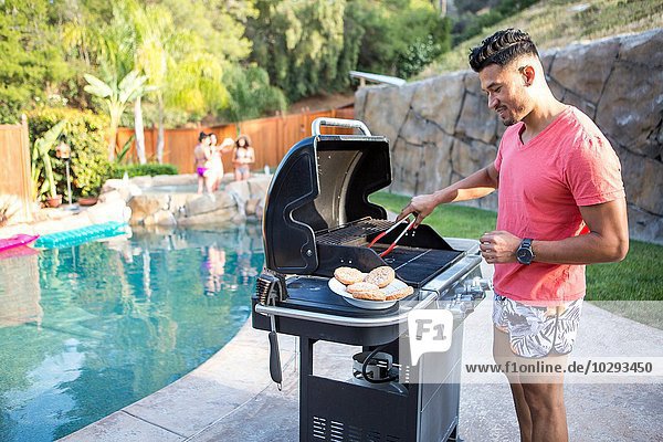 Young man cooking burgers on barbecue next to garden swimming pool