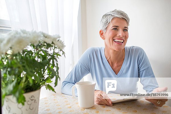 Grauhaarige reife Frau mit blauen Augen mit digitalem Tablett am Küchentisch