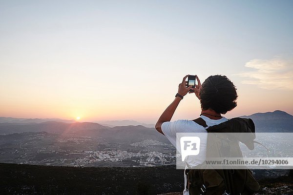 Rückansicht des jungen Mannes beim Fotografieren von Landschaft und Sonnenuntergang auf dem Smartphone  Javea  Spanien