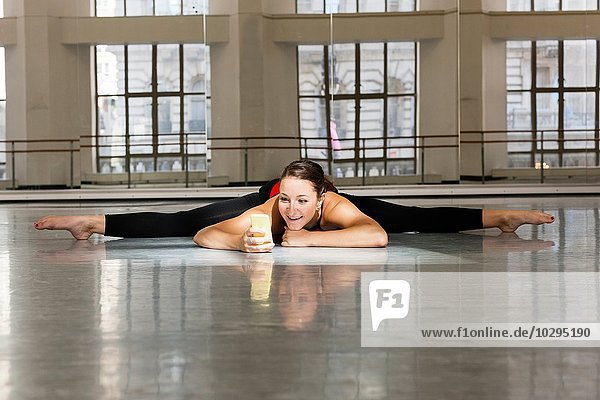 Young woman in dance studio doing the splits looking at smartphone smiling
