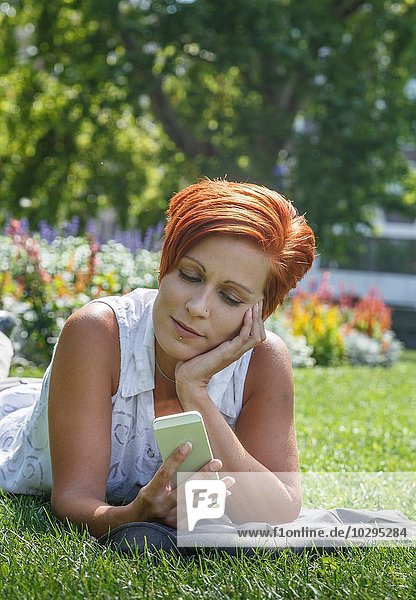 Mittlere erwachsene rothaarige Frau auf Gras auf Ellenbogen  Kinn in der Hand mit Smartphone