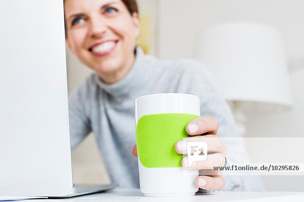 Reife Frau am Laptop hält Tasse lächelnd  Fokus auf Vordergrund
