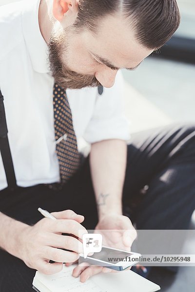 Geschäftsmann sitzt im Kreuzbein und macht Tagebuchnotizen vom Smartphone außerhalb des Büros.