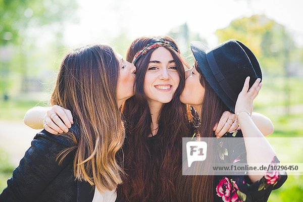 Porträt von drei stilvollen jungen Freundinnen  die sich im Park auf die Wange küssen.