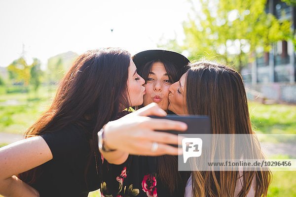 Three young female friends cheek kissing for smartphone selfie in park