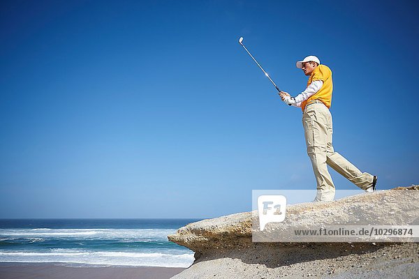 Blick auf den Golfspieler  der auf einer Klippe mit Blick auf den Ozean steht und den Golfschläger hält.