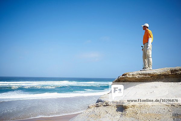 Niedriger Blickwinkel auf den Golfspieler  der auf einer Klippe mit Blick auf den Ozean steht.