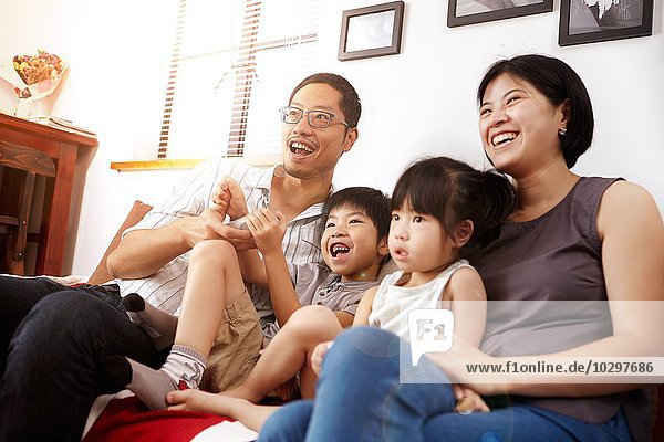 Junge  moderne chinesische Familie von Eltern und zwei kleinen Kindern  die gemeinsam zu Hause auf dem Sofa sitzen und fernsehen.