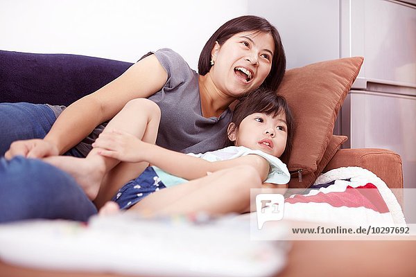 Junge chinesische Mutter und Tochter liegen auf dem Sofa und schauen gemeinsam zu Hause fernsehen.