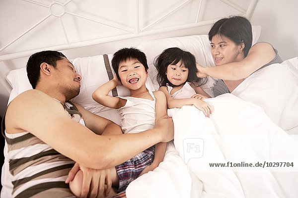 Young Chinese family of parents and two young children laying in bed together at home