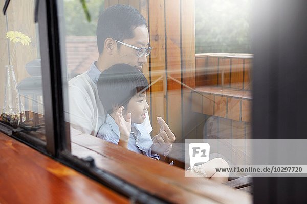 Chinesischer Vater und junger Sohn beim gemeinsamen Frühstück auf dem Balkon im Sonnenschein  durchs Fenster geschossen