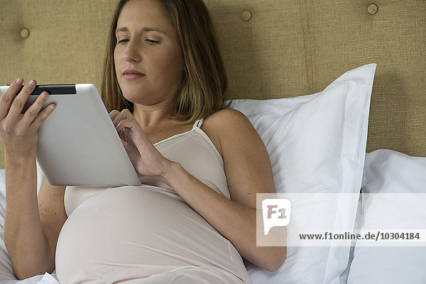 Schwangere Frau im Bett liegend mit digitalem Tablett