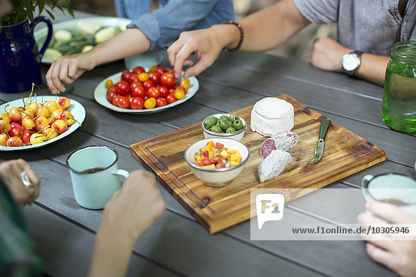 Eine Gruppe von Menschen versammelte sich um einen Tisch mit Tellern mit frischem Obst und Gemüse und einem runden Käse und Salami auf einem Schneidebrett.