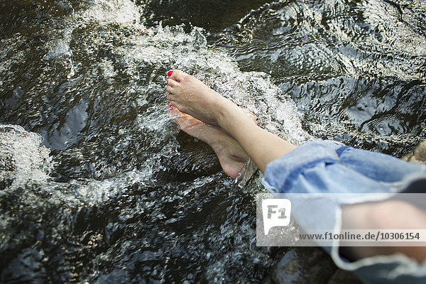 Eine Frau in modischen Jeans mit einem Riss  mit ihren Füßen im kühlen fließenden Wasser eines Flusses.
