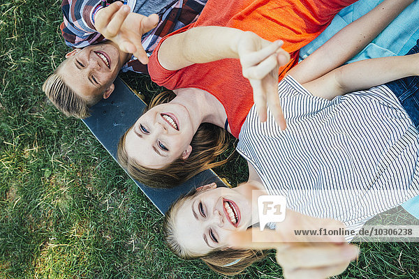 Drei glückliche Teenager-Freunde liegen auf dem Skateboard auf der Wiese und machen ein Siegeszeichen.