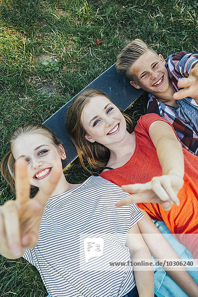Three happy teenage friends lying on skateboard in meadow making victory sign